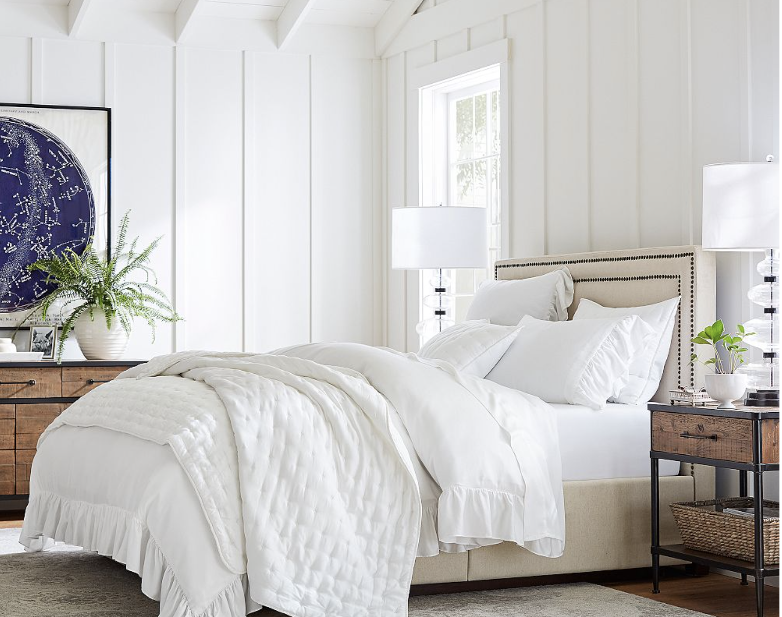 A bright bedroom featuring a soft white bed with pillows and a quilted blanket. Two wooden nightstands with lamps flank the bed, ready for your Saturday shopping finds. A large circular artwork and a potted plant adorn the left side, while a window invites natural light in.